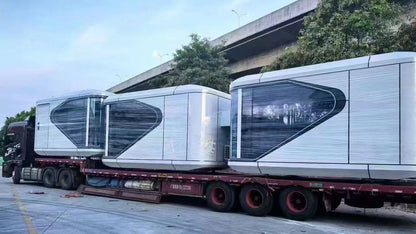 Two futuristic-looking SX-1 Lite space capsule pods on a truck with a building and trees in the background.