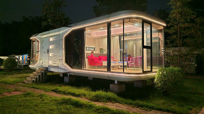 Modern prefabricated SX-2 Flow Space capsule with illuminated interior at night, surrounded by greenery.